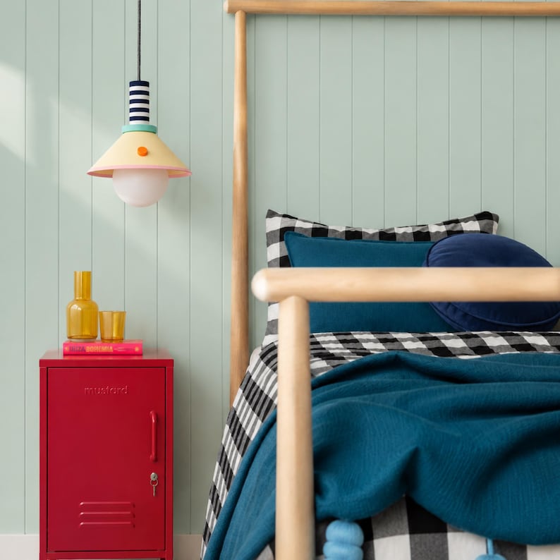 Op de afbeelding: Een rood metalen nachtkastje in locker-stijl met een deur en handgreep. Bovenop staan een gele glazen fles, een klein geel glas en een roze boek. Erboven hangt een hanglamp met een gestreepte hals en een gele, kegelvormige lampenkap.