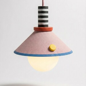 May include: A modern pendant light with a pink shade, blue trim, and a yellow button. The light fixture has a striped black and white cylinder above a red disc. A spherical, glowing light bulb hangs below.