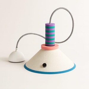 Op de afbeelding: Een hanglamp met een brede, gebroken witte conische kap met een blauwe onderrand en een kleine zwarte ronde knop. Boven de kap bevindt zich een perzikkleurige schijf en een cilinder met paarse en groenblauwe horizontale strepen aan een grijs snoer.