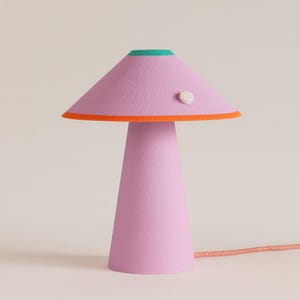 May include: A pink table lamp with a conical base and a mushroom-shaped shade. The shade has orange and green trim. A small, white button is on the side of the shade. The lamp has a textured surface and a pink cord. The lamp is a decorative lighting fixture.