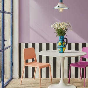 Op de afbeelding: Een witte ronde tafel op een voetstuk met een blauwe, gele en zwarte keramische kan gevuld met witte madeliefjes. De tafel wordt geflankeerd door een perzikkleurige stoel en een paarse stoel, met een hanglamp boven de tafel.