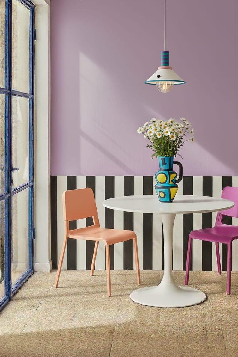Op de afbeelding: Een witte ronde tafel op een voetstuk met een blauwe, gele en zwarte keramische kan gevuld met witte madeliefjes. De tafel wordt geflankeerd door een perzikkleurige stoel en een paarse stoel, met een hanglamp boven de tafel.
