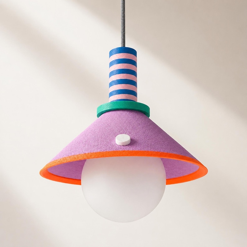 Op de afbeelding: Een hanglamp met een conische paarse kap met een feloranje rand en een klein wit rond accent. Het bovenste cilindrische gedeelte heeft blauwe en roze horizontale strepen, gescheiden door een groene ring, hangend aan een snoer.