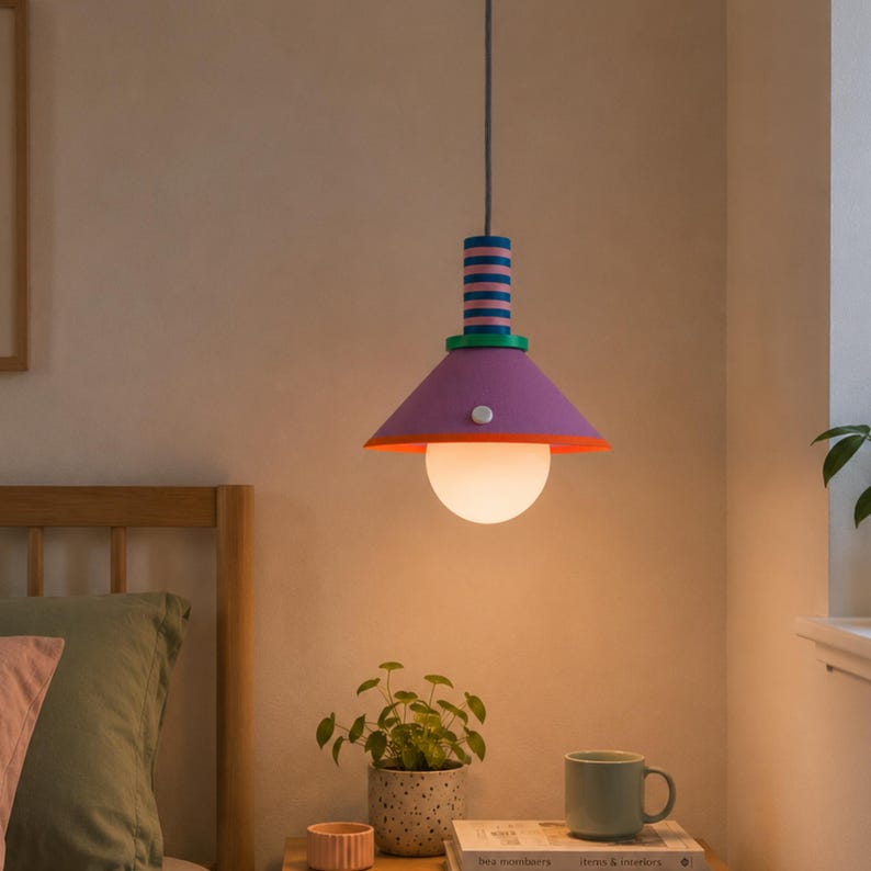 Op de afbeelding: Een hangende pendellamp met een conische paarse kap met een oranje onderkant, een groene accentring en een cilindrisch bovendeel met horizontale blauwe en roze strepen. Een bolvormige witte glazen lamp hangt onder de kap.