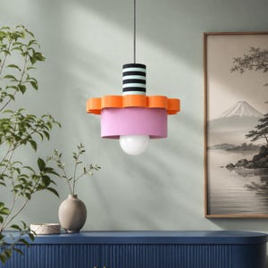 May include: A pendant light fixture featuring a cylindrical top section with black and white horizontal stripes, a scalloped orange middle tier, and a wide, smooth pink lower shade with a single round white light bulb hanging from the center.