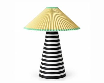 Op de afbeelding: Een tafellamp met een conische voet met horizontale zwarte en witte strepen. De lamp heeft een brede, geplooide lampenkap in een lichtgele kleur, afgewerkt met een dunne mintgroene rand langs de boven- en onderkant.