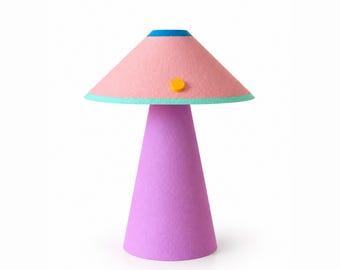 Op de afbeelding: Een tafellamp met een conische paarse voet en een brede, conische roze lampenkap. De kap heeft een felblauwe bovenrand, een groenblauwe onderrand en een kleine, ronde gele accentknop aan de zijkant van het roze oppervlak.