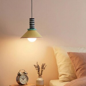 Op de afbeelding: Een hanglamp met een conische gele kap, een blauwgroene accentring en een cilindrisch bovendeel met zwarte en witte horizontale strepen. De lamp hangt aan een snoer en verspreidt een warm, gloeiend licht vanuit een ronde lamp.