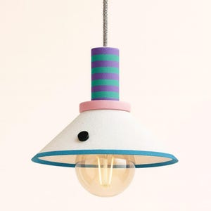 Op de afbeelding: Een hanglamp met een conische witte kap met een blauwgroene rand en een klein zwart rond accent. Het cilindrische bovendeel heeft horizontale paarse en blauwgroene strepen, gescheiden van de kap door een lichtroze ring.