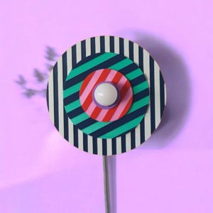 Op de afbeelding: Een kleurrijk, cirkelvormig wandkunstwerk met concentrische cirkels. De buitenste cirkel heeft marineblauwe en witte strepen, terwijl de binnenste cirkels teal, roze en rood gestreept zijn. In het midden bevindt zich een wit, bolvormig licht met een zilveren steel.