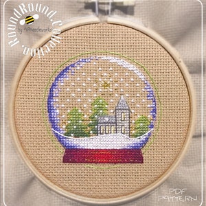 Op de afbeelding: Een kruissteekpatroon van een sneeuwbol met een kerk, bomen en sneeuwvlokken erin. De sneeuwbol staat op een rode basis en is omgeven door een groene cirkel. Het patroon zit in een houten borduurring. De tekst "PDF PATTERN" staat onderaan de afbeelding.