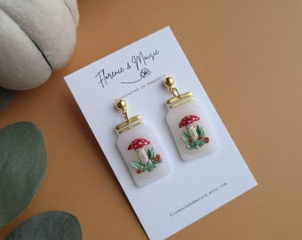 May include: White jar-shaped earrings featuring a red mushroom design, with gold-coloured details. The earrings are presented on a white card with the text "Florence & Maisie" and "Handcrafted in Hampshire".