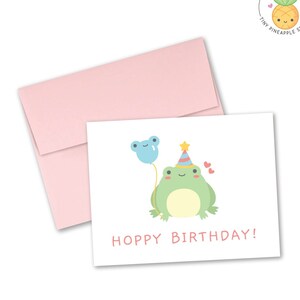 May include: A white card with a green frog wearing a party hat and holding a blue balloon. The card says "Hoppy Birthday!" in pink letters.