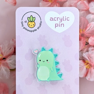 May include: A green dinosaur with a pink heart on its head, holding a pink heart in its hand. The dinosaur is an acrylic pin with the text "acrylic pin" on a purple background.