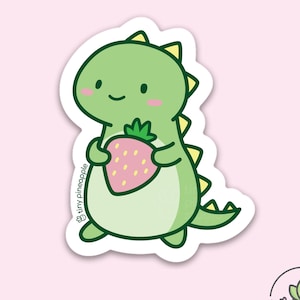 May include: A die-cut sticker featuring a cute green dinosaur holding a pink strawberry. The dinosaur has a friendly expression with rosy cheeks. The sticker has a white border and the text "tiny pineapple studio".