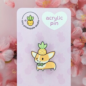 May include: A light pink card with the text "acrylic pin" in a white heart shape. The card features a cartoon image of a corgi wearing a pineapple on its head. The card also has a logo for "tiny pineapple studio" with a small pineapple graphic.