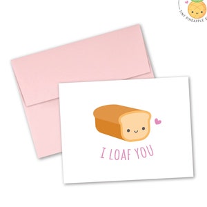 May include: A white card with a pink envelope. The card has a cartoon illustration of a loaf of bread with a smiling face and the text "I LOAF YOU".