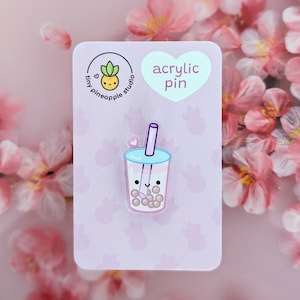 May include: A clear acrylic pin featuring a cute cartoon boba tea drink with a pink and white color scheme. The pin has a light blue lid and straw, with a small pink heart. The pin is on a pink backing card with the text "acrylic pin".
