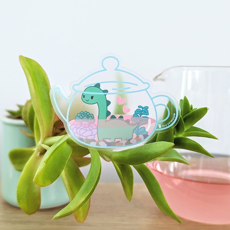 May include: A clear glass teapot with a green dinosaur inside. The teapot is filled with small plants and rocks. The sticker has a pink heart design and the text "tiny pineapple" on the side.
