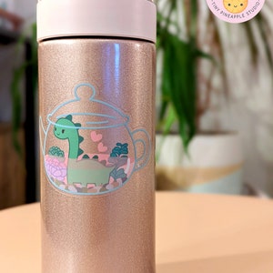 May include: A rose gold stainless steel water bottle with a sticker of a green dinosaur inside a teapot with pink hearts and green plants. The sticker is on the front of the bottle.