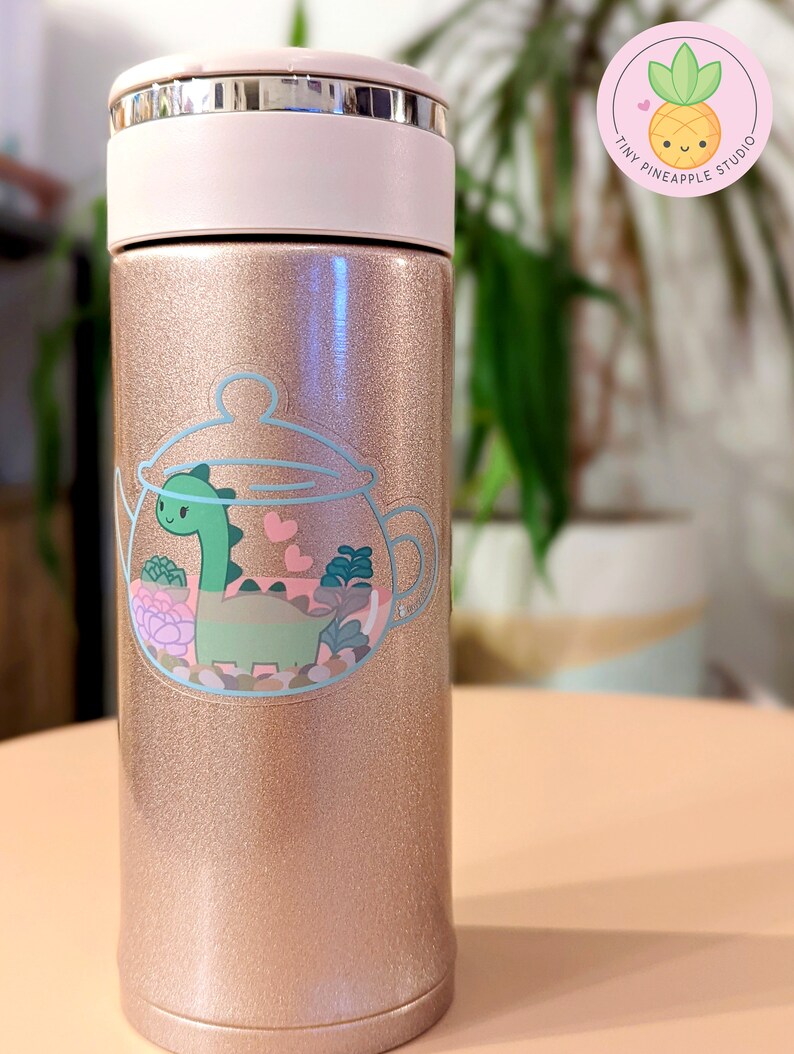 May include: A rose gold stainless steel water bottle with a sticker of a green dinosaur inside a teapot with pink hearts and green plants. The sticker is on the front of the bottle.