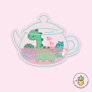 May include: A white ceramic teapot with a green dinosaur inside. The teapot is filled with pink and green succulents. The teapot has a light blue lid and handle. The text "tiny pineapple" is written on the side of the teapot.