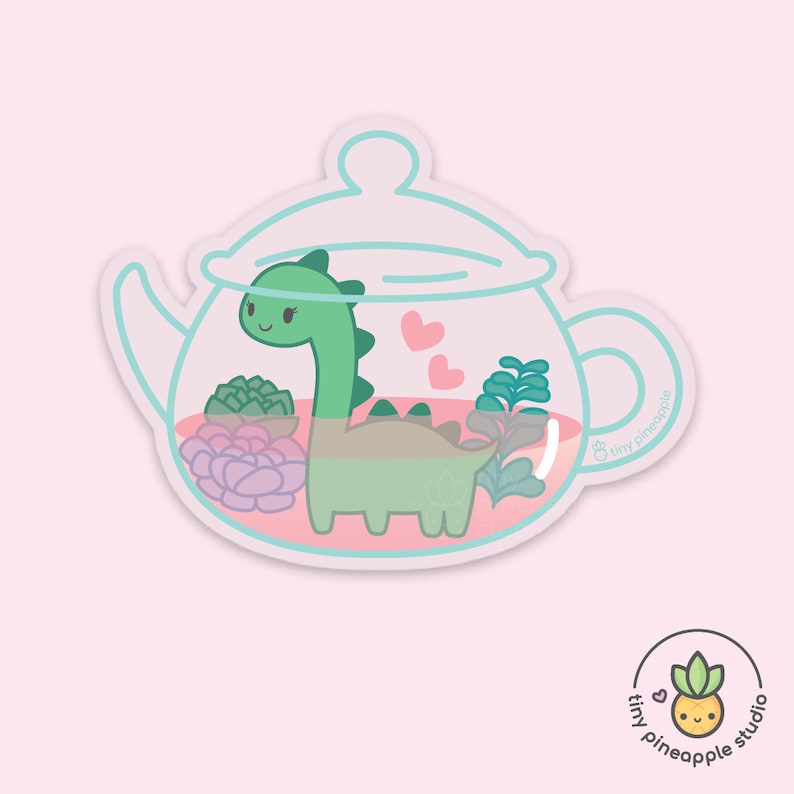 May include: A white ceramic teapot with a green dinosaur inside. The teapot is filled with pink and green succulents. The teapot has a light blue lid and handle. The text "tiny pineapple" is written on the side of the teapot.