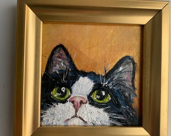 May include: An oil painting of a black and white cat with bright green eyes and a pink nose, set against a golden yellow background. The artwork is encased in a square, metallic gold frame with a beveled edge and a smooth, reflective finish.