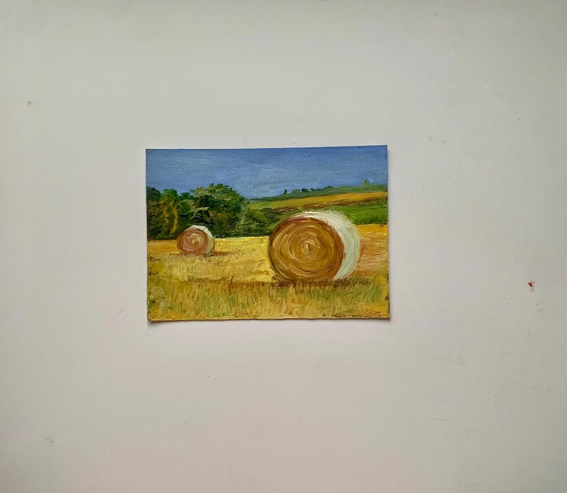 May include: Oil painting of a rural landscape featuring two hay bales in a field of golden grass. The sky is a gradient of blue, and green trees and hills are in the background. The painting has a textured, impressionistic style.