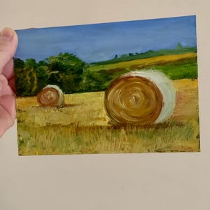 May include: An oil painting depicting a rural landscape with two hay bales in a field. The bales are brown and white, set against a backdrop of green trees, rolling hills, and a blue sky. The artwork is on a small rectangular canvas.