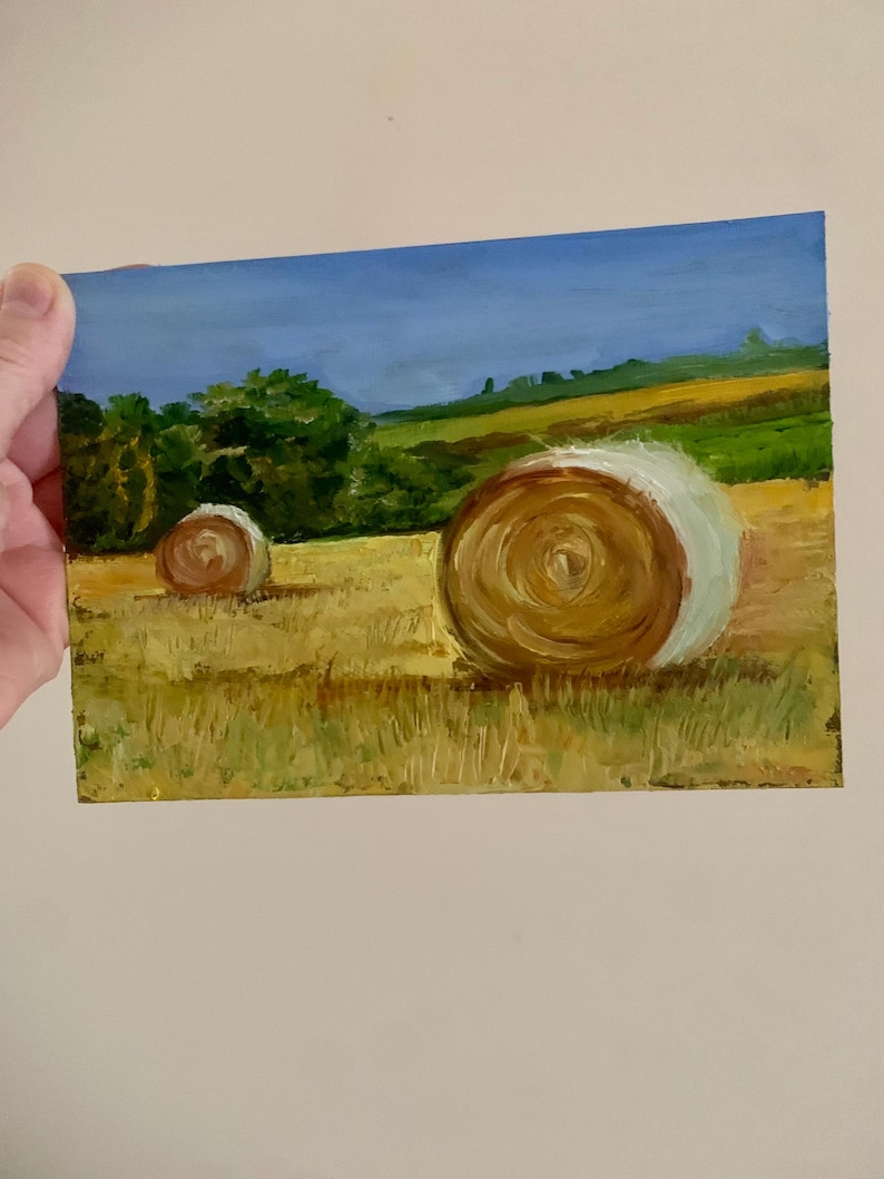 May include: An oil painting depicting a rural landscape with two hay bales in a field. The bales are brown and white, set against a backdrop of green trees, rolling hills, and a blue sky. The artwork is on a small rectangular canvas.