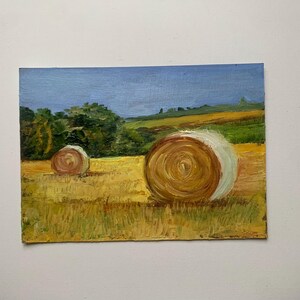 May include: Oil painting of a rural landscape featuring two hay bales in a field of golden grass. The sky is a light blue, and trees and hills are visible in the background. The artwork has a textured, impressionistic style.