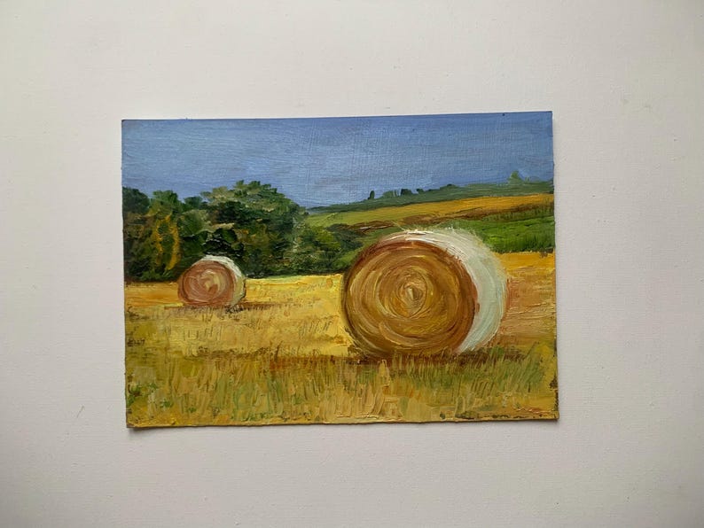 May include: Oil painting of a rural landscape featuring two hay bales in a field of golden grass. The sky is a light blue, and trees and hills are visible in the background. The artwork has a textured, impressionistic style.
