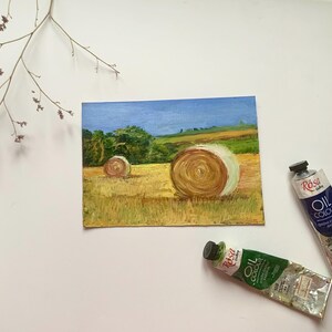 May include: An oil painting of a rural landscape featuring two hay bales in a field under a blue sky. The painting is accompanied by two tubes of oil paint. A sprig of dried flowers is in the upper left corner.