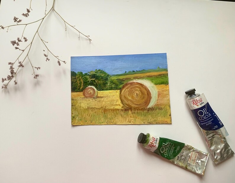 May include: An oil painting of a rural landscape featuring two hay bales in a field under a blue sky. The painting is accompanied by two tubes of oil paint. A sprig of dried flowers is in the upper left corner.