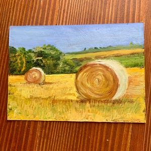 May include: Oil painting of a rural landscape featuring two hay bales in a golden field under a blue sky. Green trees and rolling hills are in the background. The artwork is on a rectangular canvas.