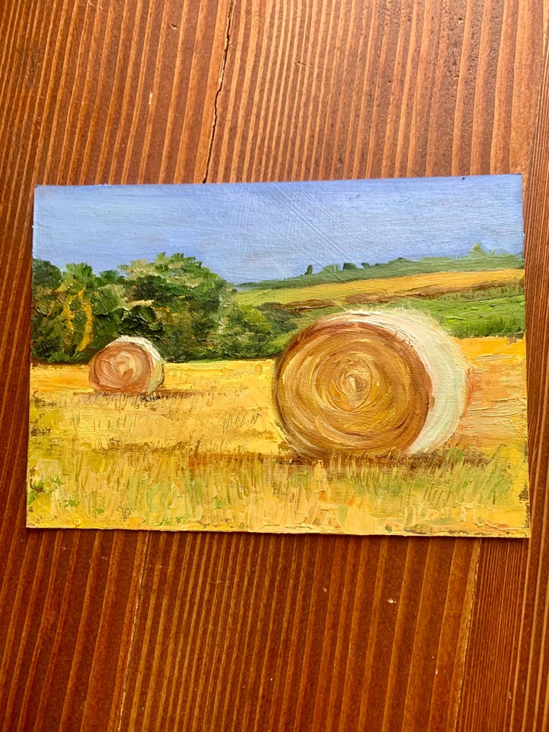 May include: Oil painting of a rural landscape featuring two hay bales in a golden field under a blue sky. Green trees and rolling hills are in the background. The artwork is on a rectangular canvas.
