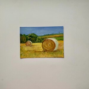 May include: Oil painting of a rural landscape featuring two large hay bales in a field of golden grass. The sky is a gradient of blue, and green trees and hills are in the background. The painting has a textured, impressionistic style.