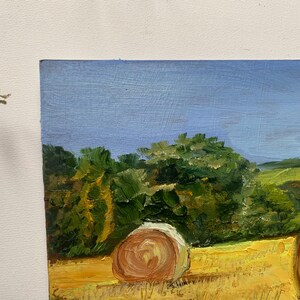 May include: Oil painting of a rural landscape featuring a field of hay bales under a blue sky. The artwork depicts a sunny day with green trees and rolling hills in the background. The painting is on canvas.