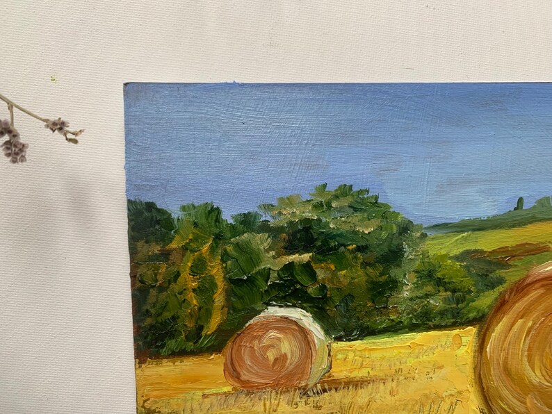 May include: Oil painting of a rural landscape featuring a field of hay bales under a blue sky. The artwork depicts a sunny day with green trees and rolling hills in the background. The painting is on canvas.