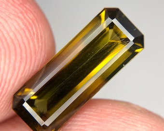 May include: A faceted, rectangular, green and brown gemstone with a light brown hue. The gemstone is cut into a rectangular shape with a slightly rounded edge.