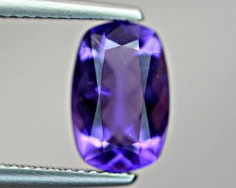 May include: A close-up of a purple gemstone with a faceted cut. The gemstone is held in a pair of tweezers.