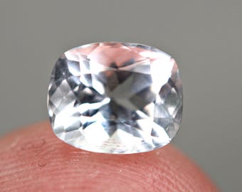 May include: A faceted, cushion-cut gemstone with a pale, translucent appearance. The gem is held between two fingers, showcasing its clarity and the play of light across its many facets. The stone appears to be a light, almost colorless, crystal.