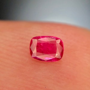May include: A vibrant, deep red gemstone with a cushion cut. The gemstone is faceted and has a glossy finish.
