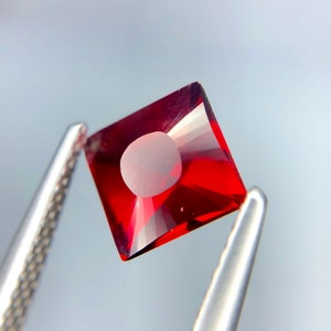 Puede incluir: Una piedra preciosa cuadrada de color rojo oscuro con un corte facetado. La piedra preciosa está sujeta por unas pinzas.