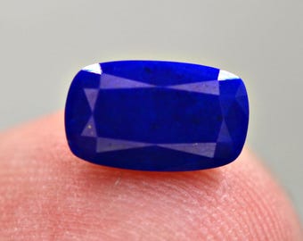 May include: A faceted, deep blue gemstone with a rectangular cushion cut. The gem's facets reflect light, creating a sparkling effect. The stone is a vibrant shade of blue, with a polished surface.