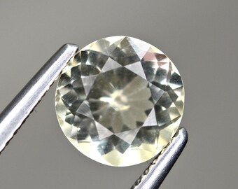 May include: A round, faceted gemstone held by silver tweezers. The gem has a pale yellow hue and a complex, geometric pattern of triangular facets. The tweezers are in the foreground, and the background is a solid, neutral gray.