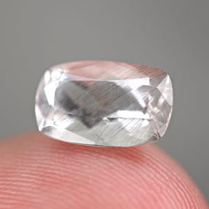 Puede incluir: Una gema clara y facetada con inclusiones internas, apoyada en la yema de un dedo. La gema tiene forma rectangular con esquinas redondeadas y una superficie pulida. Las inclusiones aparecen como finas líneas plateadas.