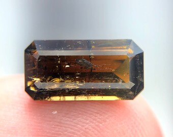 May include: A dark brown, faceted gemstone with a rectangular shape. The gemstone has a slightly cloudy appearance with visible inclusions.