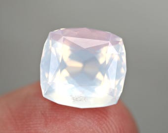 May include: A faceted, cushion-cut gemstone with a milky white and iridescent appearance. The gem is held between fingers, showcasing its polished surfaces and internal reflections. The gem's shape is square with rounded corners.